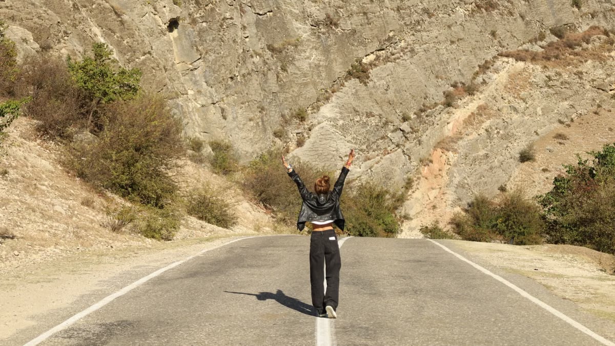 A woman in a leather jacket stands joyfully on a rural road surrounded by rocky terrain.