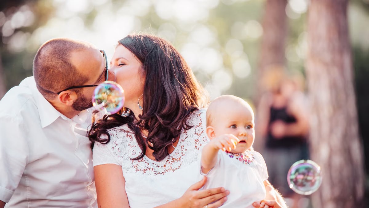 A tender family moment captured with a couple kissing and a baby holding bubbles outdoors.